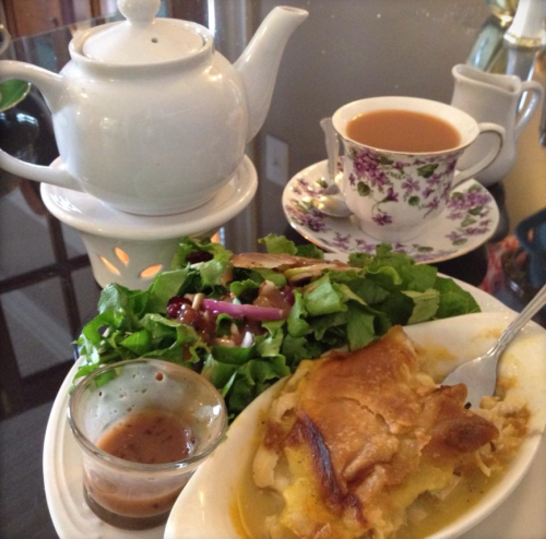A white teapot, a cup of tea, a salad, and a dish of baked casserole on a table.