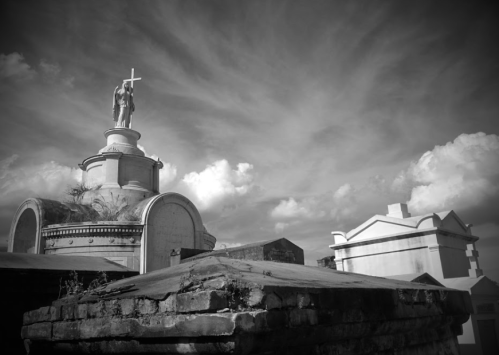 A black and white image of a cemetery with a statue of a figure on a tomb and clouds in the background.