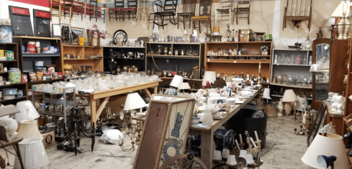 A cluttered antique shop filled with various items, including lamps, glassware, and furniture, creating a nostalgic atmosphere.