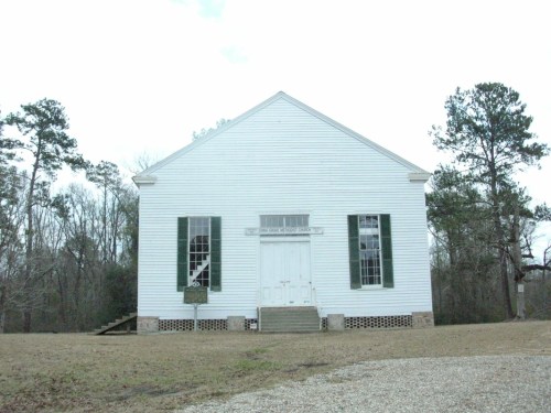 A white, historic church building with green shutters, surrounded by trees and a gravel path.