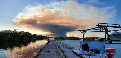 A person stands by a calm waterway, watching a large, billowing cloud against a sunset sky. A truck is parked nearby.