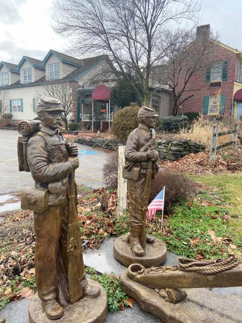 Two bronze statues of soldiers stand in a garden, with a small American flag nearby and a house in the background.