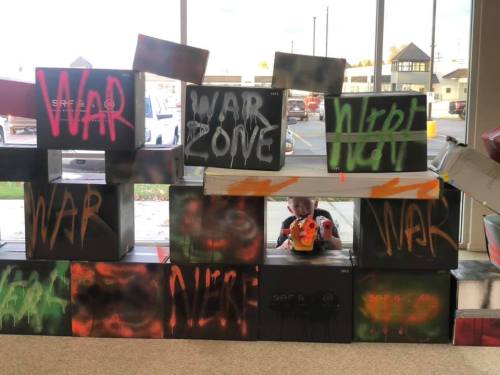 A child peeks out from behind colorful boxes labeled with graffiti-style words like "WAR" and "WAR ZONE."