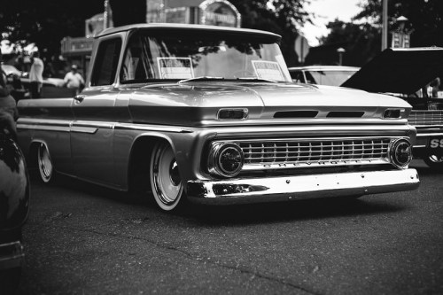 A classic silver pickup truck with a lowered stance, parked at a car show, surrounded by trees and other vehicles.
