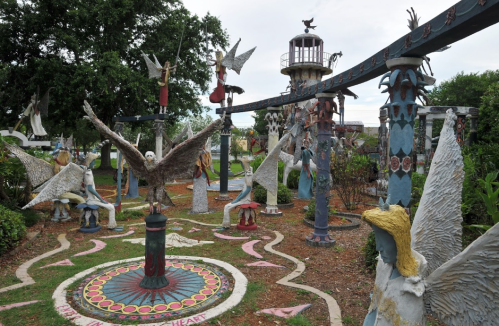 A whimsical outdoor garden featuring colorful sculptures of angels and birds, with a lighthouse in the background.
