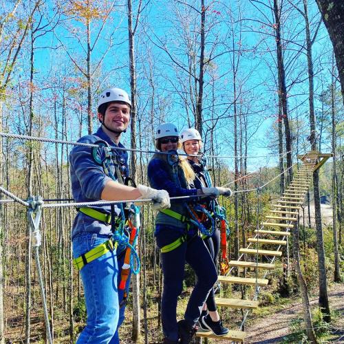 Three people in helmets and harnesses smile while standing on a zip line platform among trees on a sunny day.