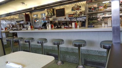 A retro diner interior featuring a counter with stools, a menu board, and a display of food items.