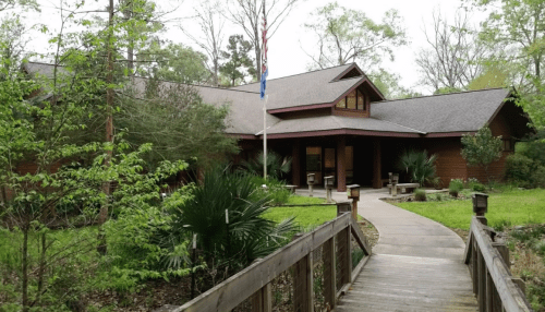 A wooden building surrounded by greenery, with a pathway leading to the entrance and an American flag in front.