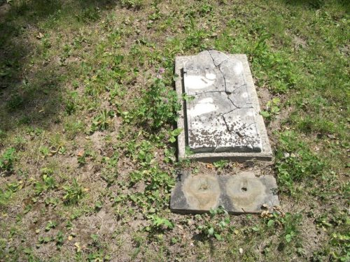 A cracked gravestone lies on the ground, surrounded by grass and small plants.