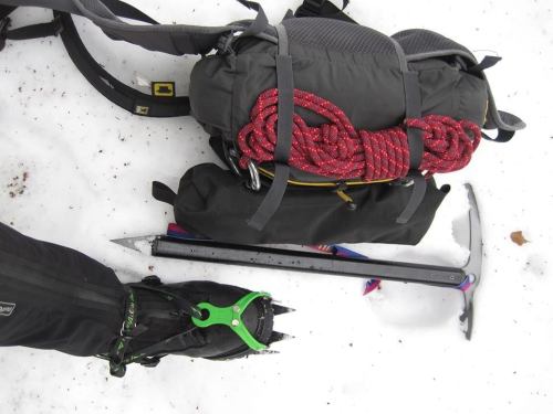 Climbing gear on snow: a backpack, climbing rope, ice axe, and crampon on a boot.