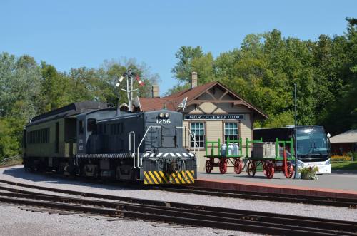 A vintage train at a station with a historic building, surrounded by trees and a parked bus nearby.