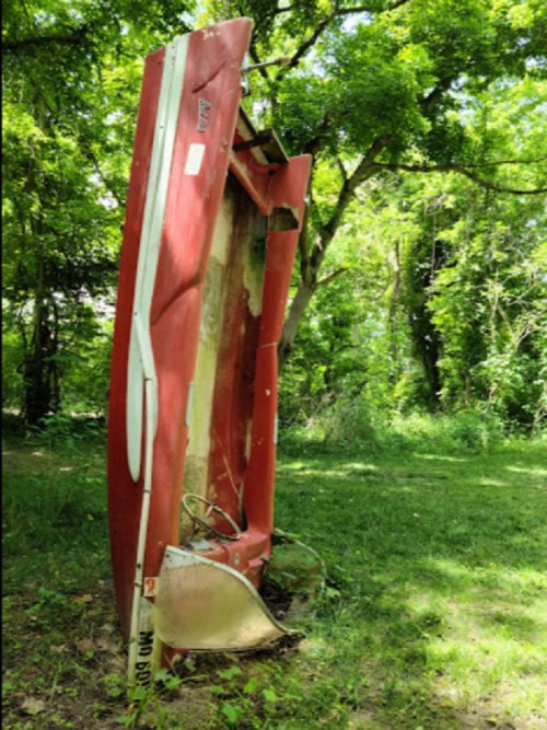 A red, overturned boat rests on its side in a grassy area surrounded by trees.