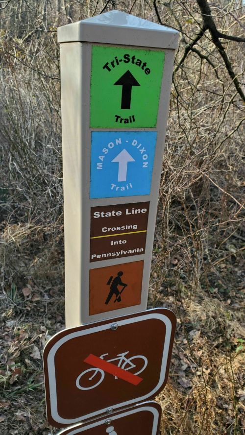 Trail sign indicating the Tri-State Trail, Mason-Dixon Trail, and a state line crossing into Pennsylvania. No biking allowed.