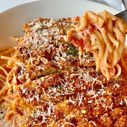 A fork holding spaghetti with crispy breaded chicken, topped with cheese and herbs in a white bowl.