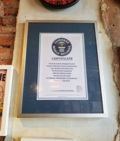 Framed Guinness World Records certificate for the largest collection of pizza-related items, displayed on a brick wall.