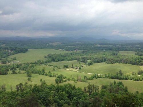A panoramic view of lush green fields and rolling hills under a cloudy sky. Mountains are visible in the distance.