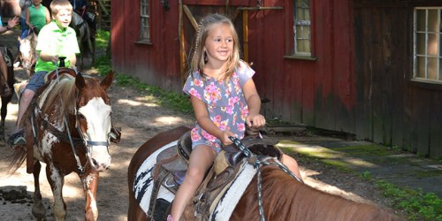 A young girl in a floral outfit smiles while riding a horse on a trail, with other riders in the background.