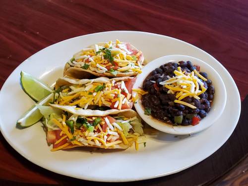Three tacos filled with lettuce, tomatoes, and cheese, served with black beans and lime wedges on a white plate.