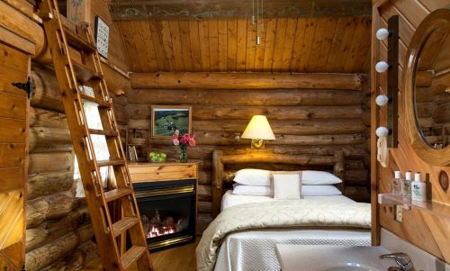 Cozy log cabin interior featuring a bed, fireplace, wooden ladder, and rustic decor. Warm and inviting atmosphere.