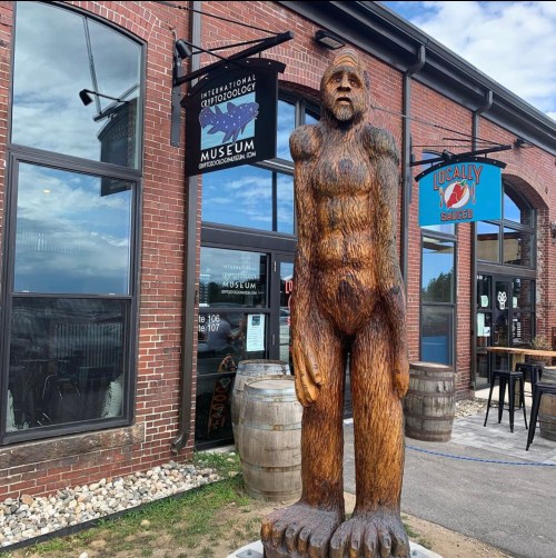 A large wooden Bigfoot statue stands outside the International Cryptozoology Museum, with brick walls and barrels nearby.