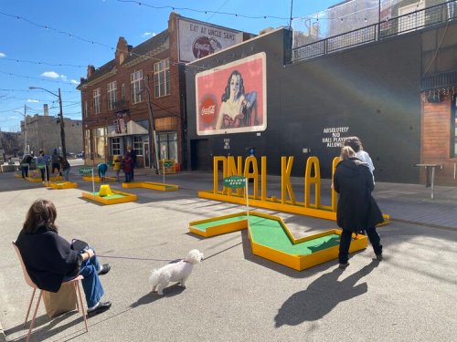 A street scene with colorful mini-golf setups, people playing, and a vintage Coca-Cola sign in the background.