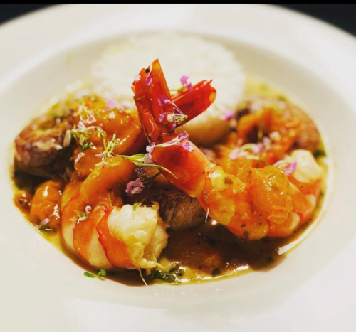 A gourmet dish featuring shrimp, rice, and garnishes, served in a rich sauce with colorful herbs and flowers.