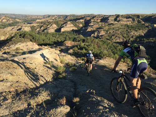 Two mountain bikers navigate a rocky trail overlooking a vast, rugged landscape under a clear blue sky.