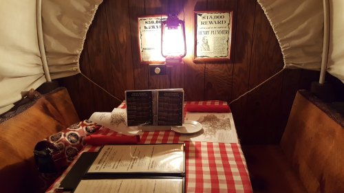 Cozy dining area inside a covered wagon, featuring a checkered tablecloth, menus, and vintage lanterns on the walls.