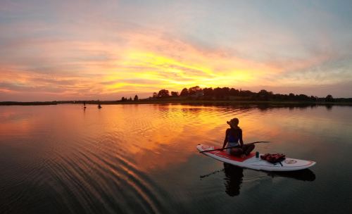 A person paddles on a stand-up paddleboard at sunset, reflecting vibrant colors on calm water.