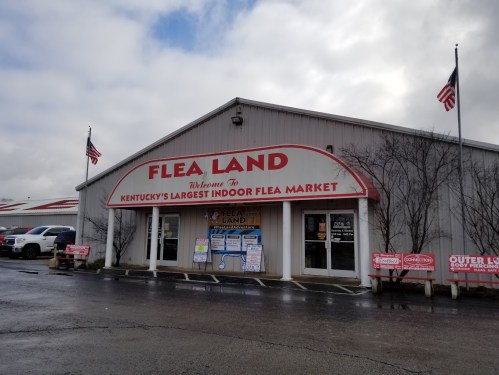 Sign for "Flea Land," Kentucky's largest indoor flea market, with flags and a cloudy sky in the background.