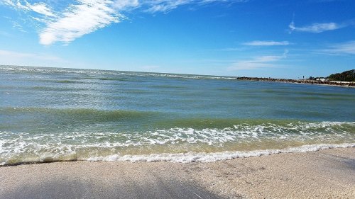 A serene beach scene with gentle waves, clear blue skies, and a distant shoreline under a few wispy clouds.