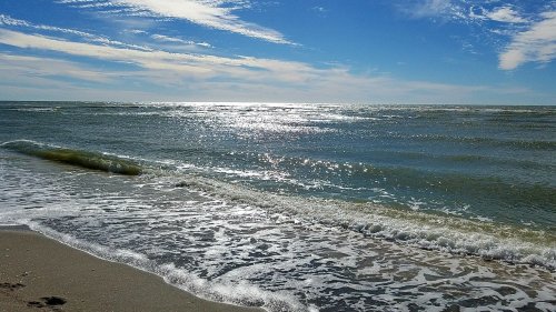 A serene beach scene with gentle waves lapping at the shore under a bright blue sky and scattered clouds.