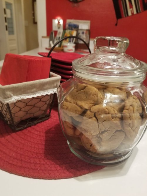 A glass jar filled with cookies sits on a table next to a basket of napkins and red plates.
