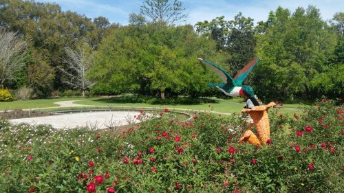 A colorful bird sculpture perched on a pedestal surrounded by blooming roses in a lush garden.