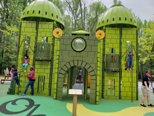 Children play on a vibrant green playground structure surrounded by trees, with some climbing and others exploring.