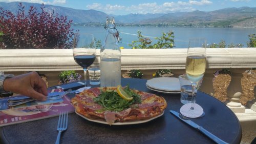 A scenic dining table with a pizza topped with arugula and prosciutto, wine glasses, and a view of a lake and mountains.