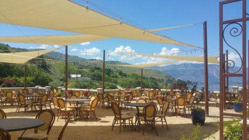 Outdoor dining area with shaded tables and chairs, overlooking green hills and a lake under a blue sky with clouds.