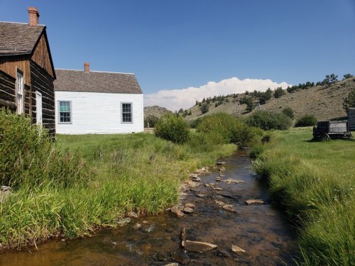 A serene landscape featuring a stream, grassy areas, and two rustic buildings under a clear blue sky.