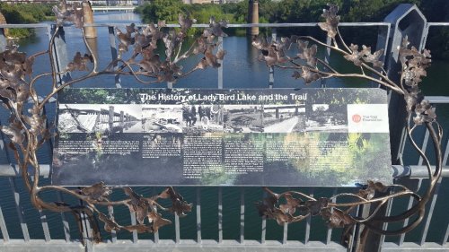 Sign about the history of Lady Bird Lake and the trail, surrounded by decorative metal foliage, with a river in the background.