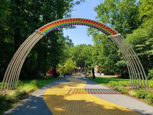 A colorful rainbow arch over a winding yellow brick path in a lush green park with playgrounds in the background.