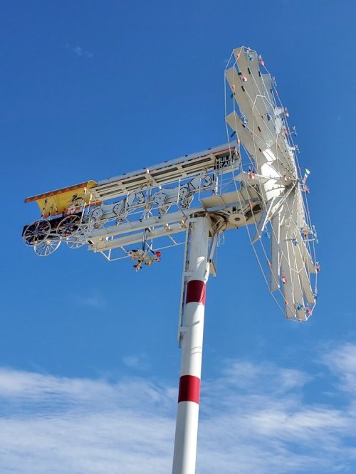 A large, colorful windmill structure with a vintage cart attached, set against a clear blue sky.