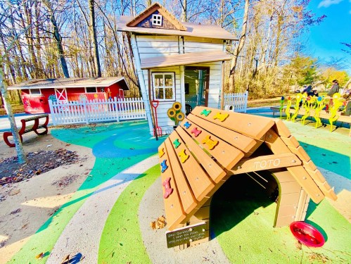 A colorful playground featuring a small house, a doghouse with climbing toys, and a red barn in a wooded area.