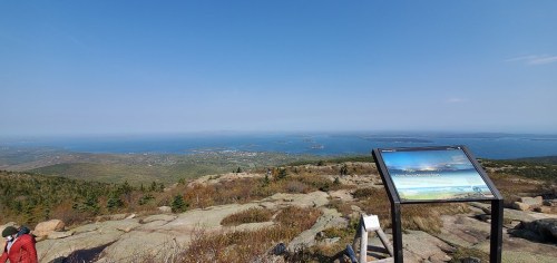 A scenic view from a rocky overlook, featuring a sign and a distant coastline under a clear blue sky.