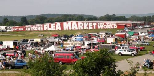 A bustling flea market with numerous vendors, cars, and a large sign reading "The Most Awesome Flea Market in the World."
