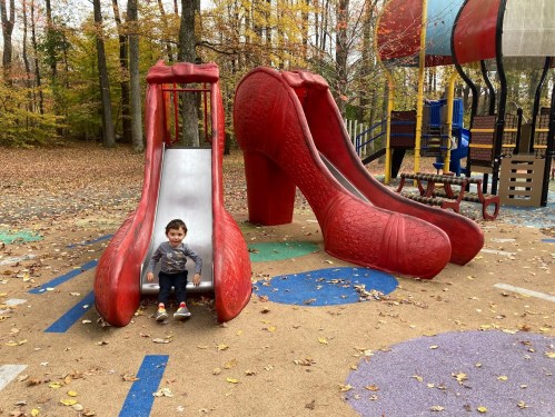 A child sits at the base of a large red high-heeled shoe slide in a colorful playground surrounded by autumn leaves.