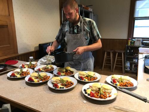A chef plates multiple dishes of eggs Benedict with vegetables and fruit on a kitchen counter.