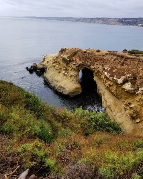 A coastal cliff with a natural arch, overlooking calm waters and distant hills under a cloudy sky.