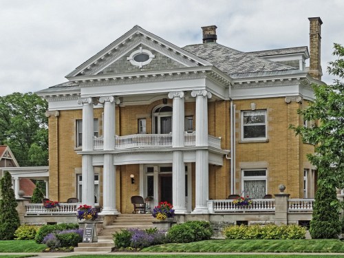 A grand two-story yellow brick house with white columns, a balcony, and colorful flower beds in front.
