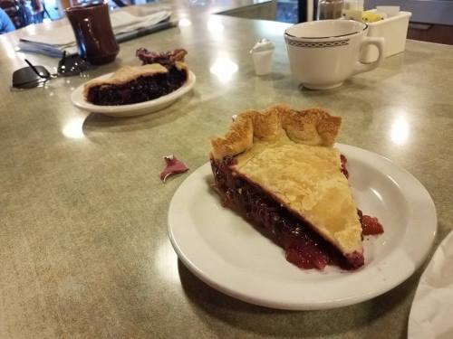 Two slices of berry pie on white plates, with a coffee cup and condiments in the background on a diner table.