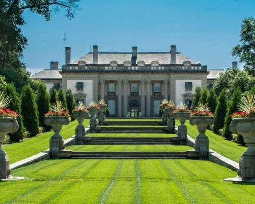 A grand mansion surrounded by manicured lawns and decorative flower pots, under a clear blue sky.